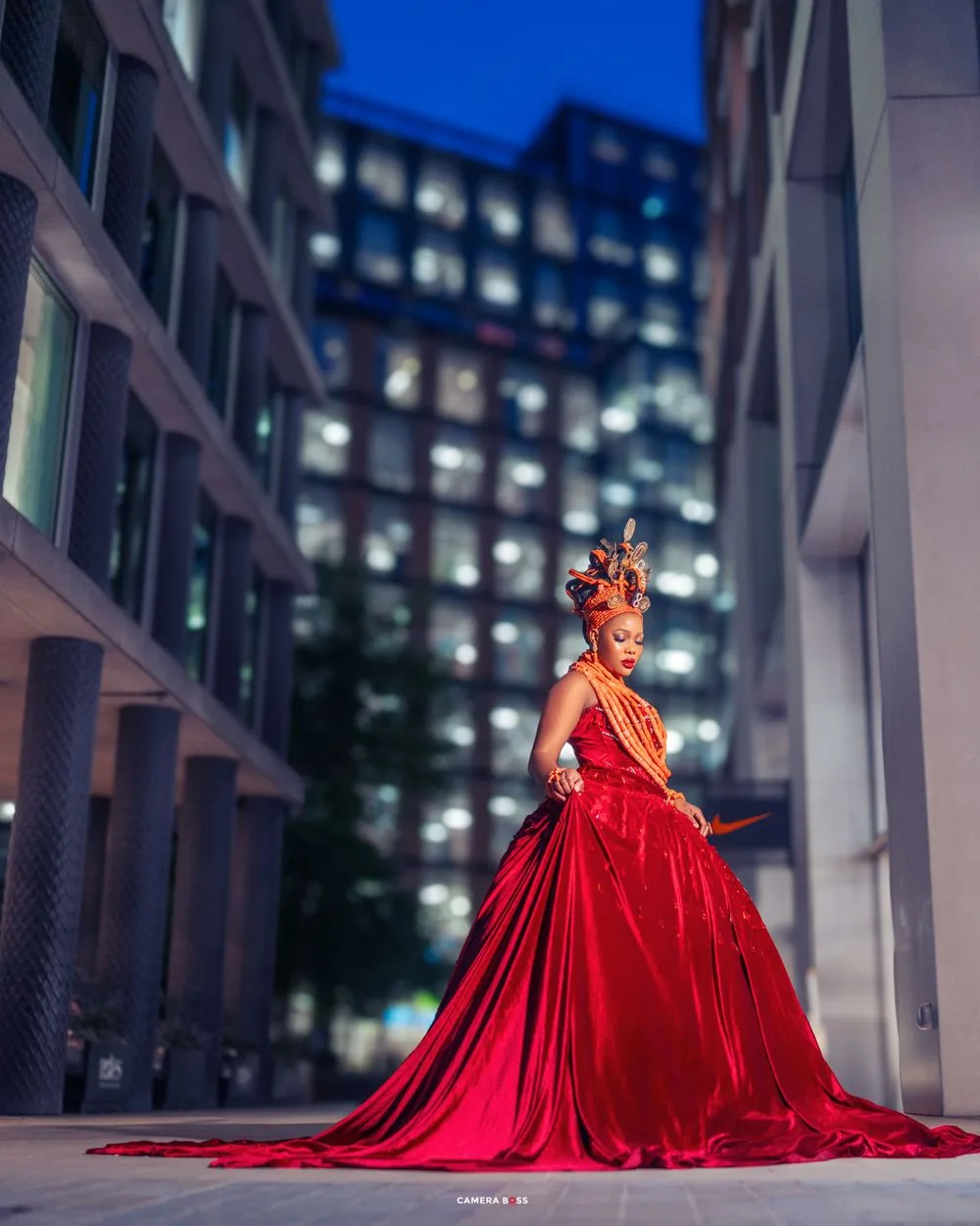 A stunning red ball gown with dramatic train photographed against illuminated city buildings at dusk.