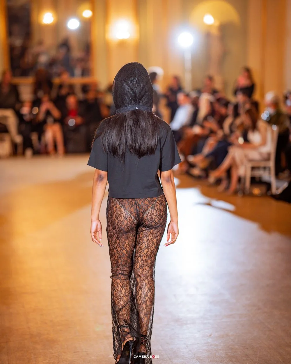 Fashion model wearing black top and lace pants walks runway at elegant fashion show with chandeliers.