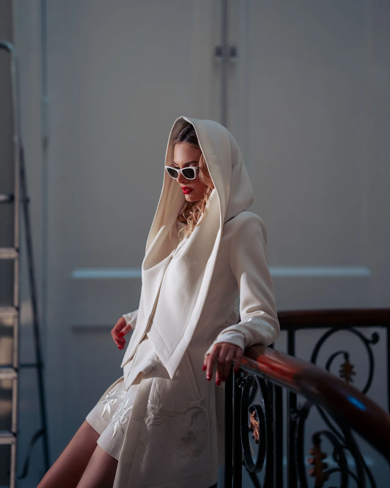 Fashion model in white hooded dress and sunglasses poses elegantly on ornate staircase railing.