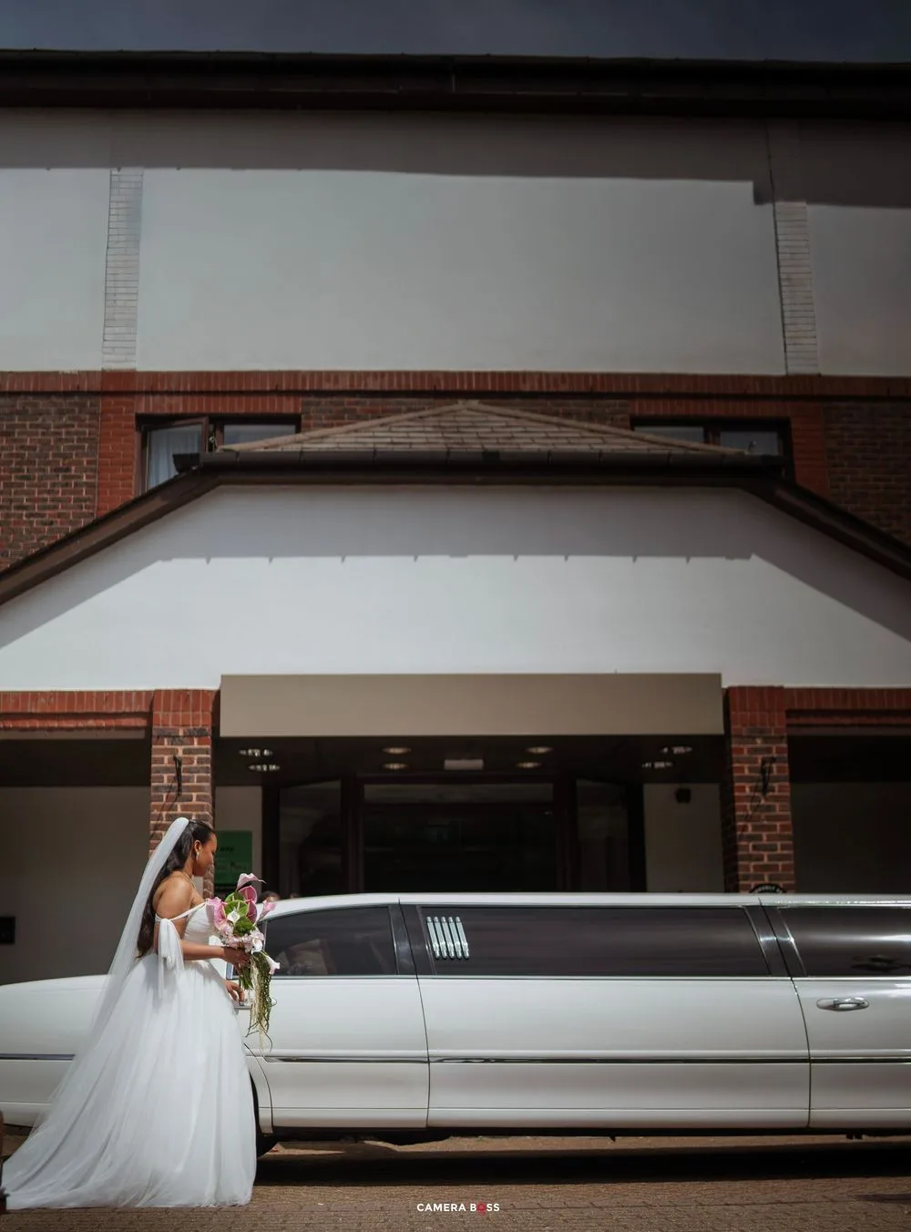Bride in white gown and veil holding bouquet beside a white stretch limousine outside a brick building.