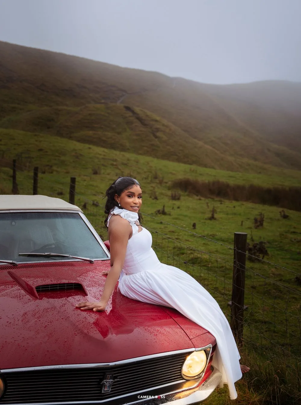 Woman in white dress posed on red classic car hood with misty green hills in background.