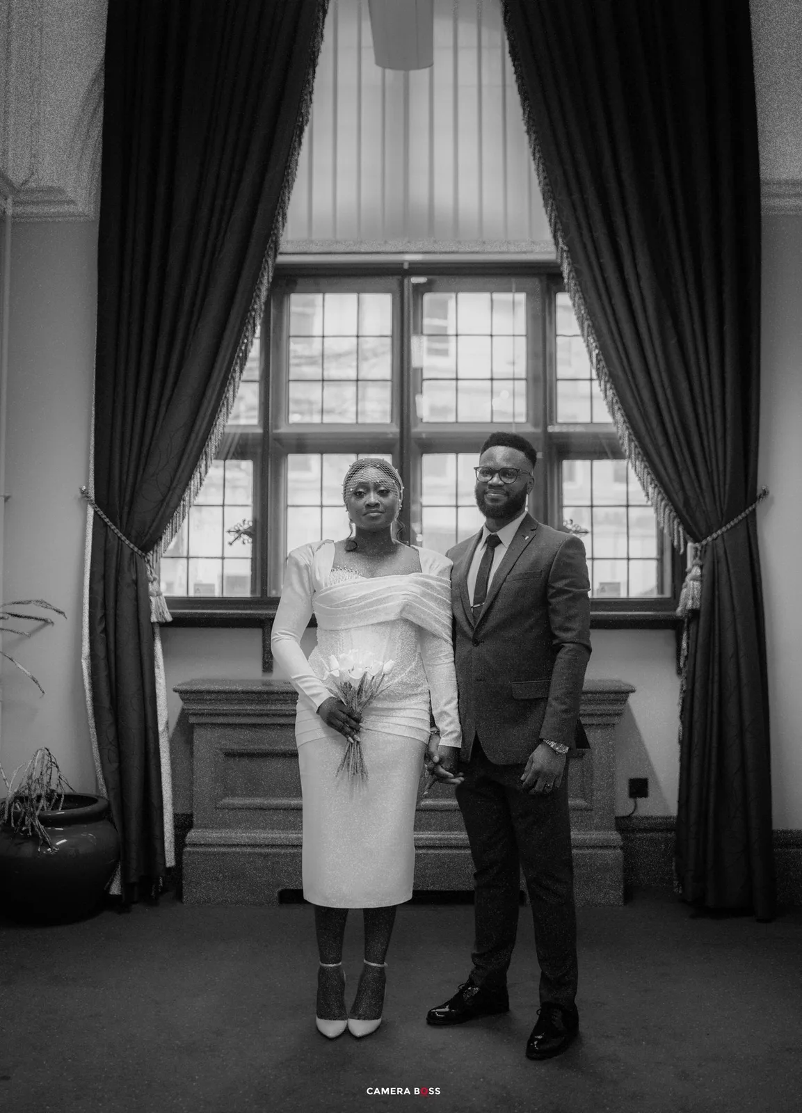 A black and white wedding photograph showing a couple in elegant attire standing in front of a large window with dark curtains.