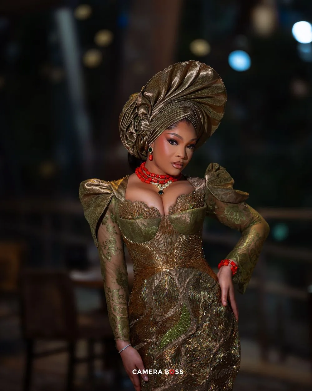 A model in an ornate gold and green baroque-style gown with a dramatic headpiece poses against a dark background.