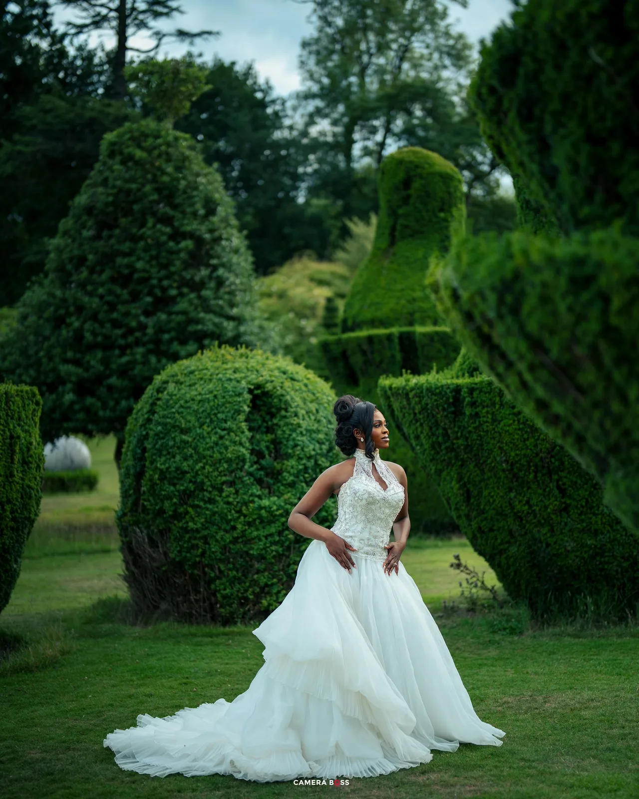 Elegant white bridal gowns are displayed amidst lush garden landscaping with manicured hedges and topiary.