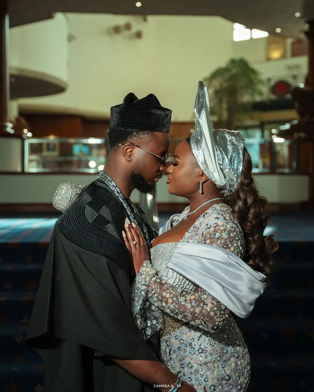 Couple in elegant Nigerian traditional attire sharing a romantic moment, man in black agbada, woman in silver gele headwrap.