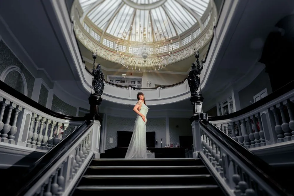 Elegant wedding dress silhouette on grand staircase beneath ornate domed skylight in historic mansion interior.