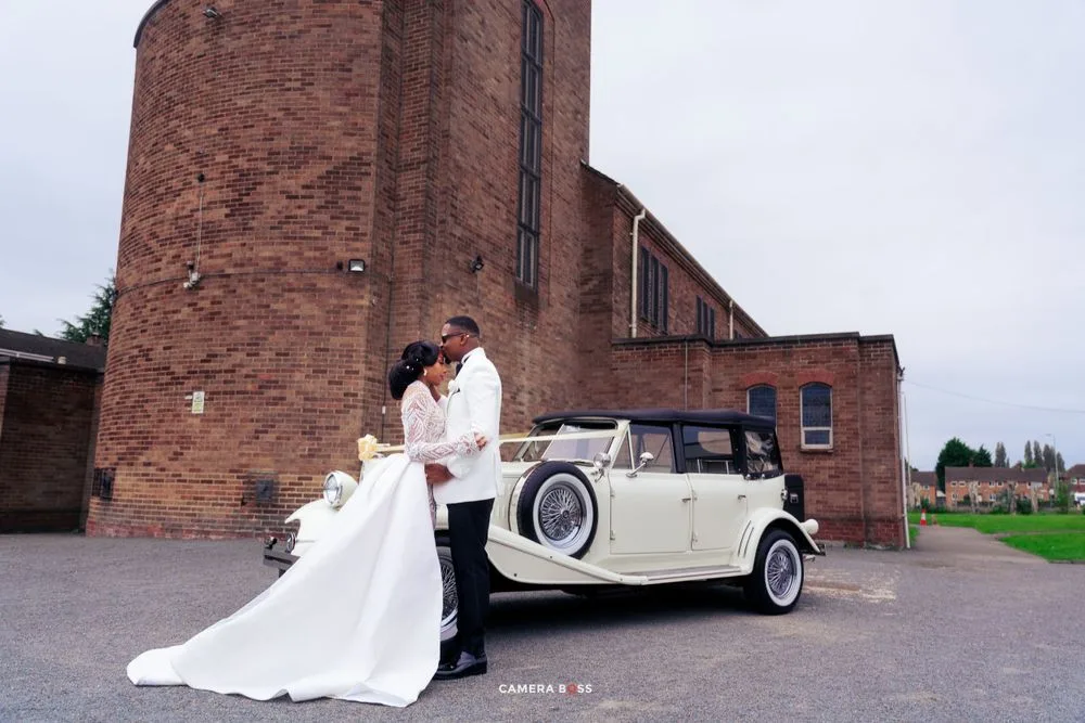 A vintage cream-colored Rolls Royce parked in front of a brick church serves as backdrop for a romantic wedding photo.