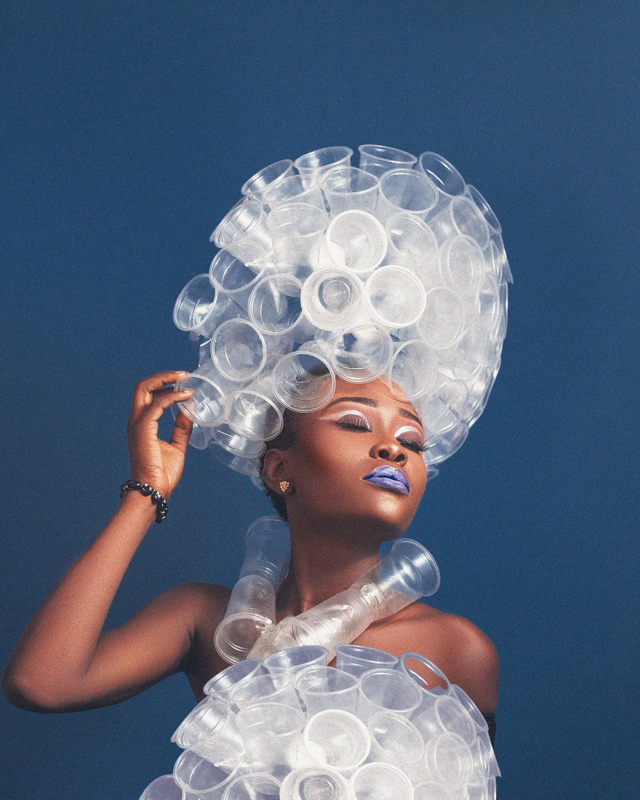 Creative fashion portrait featuring clear plastic cup accessories arranged in a sculptural headpiece and neckpiece against blue.