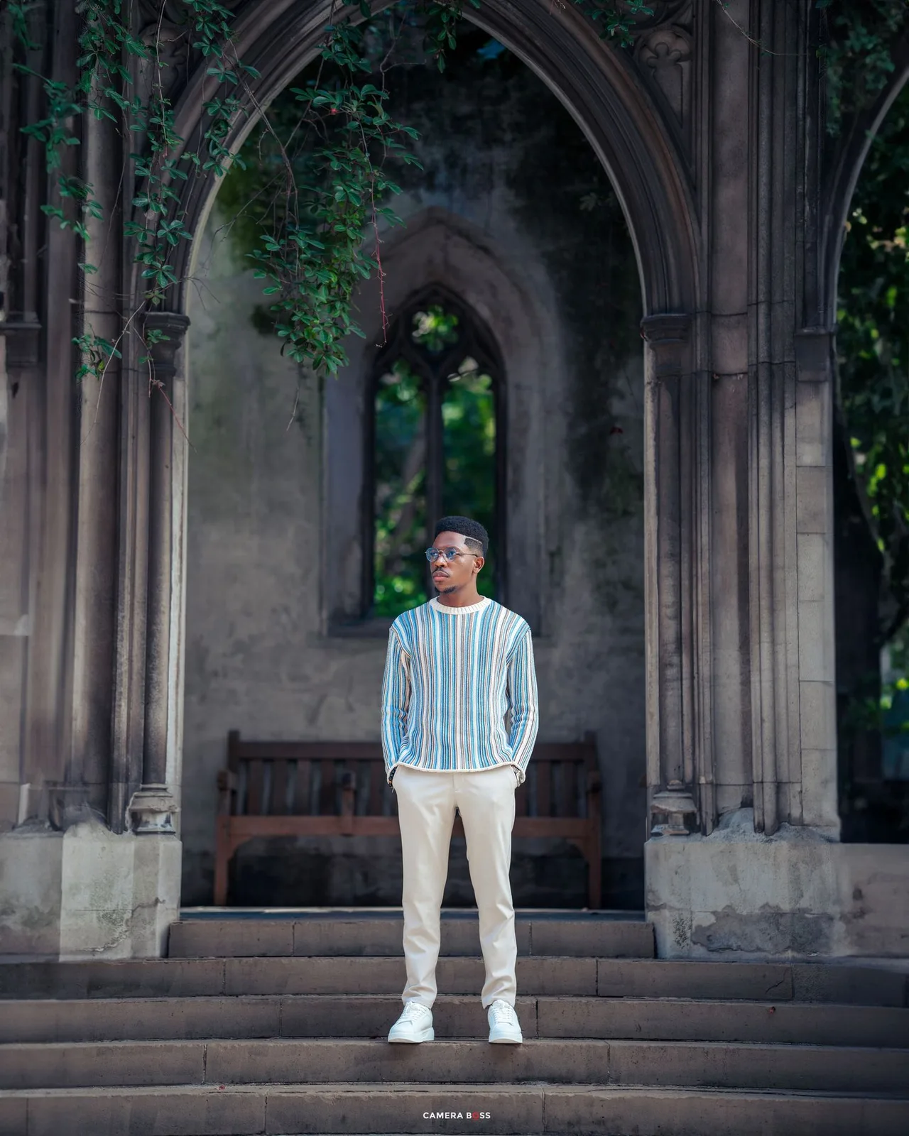 A figure stands in a striped blue and white sweater under a Gothic stone archway with a bench visible in the background.