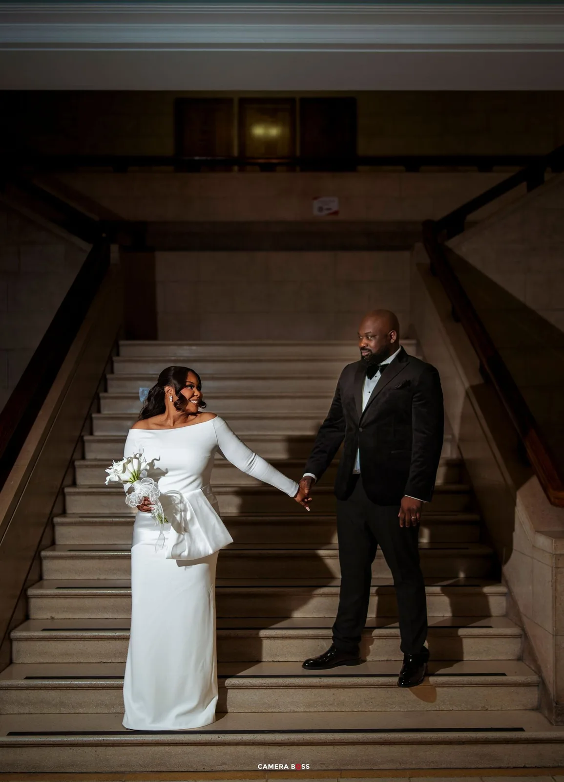 Bride in white peplum gown and groom in black tuxedo hold hands on grand staircase at elegant wedding venue.