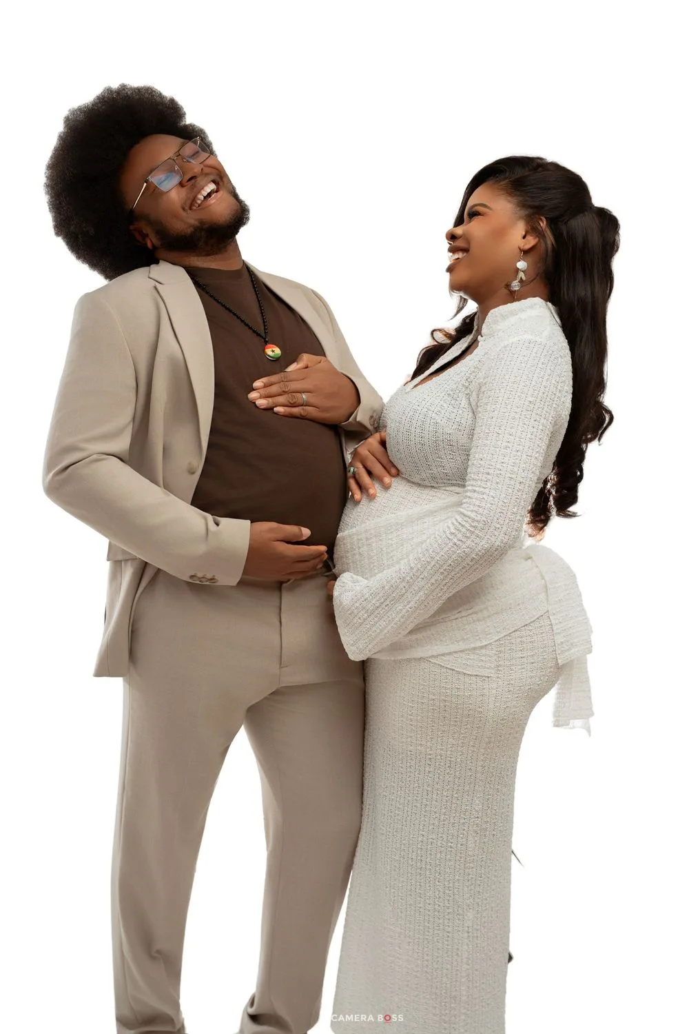 A joyful pregnant couple poses together, man in beige suit and woman in white dress, both smiling and cradling her baby bump.