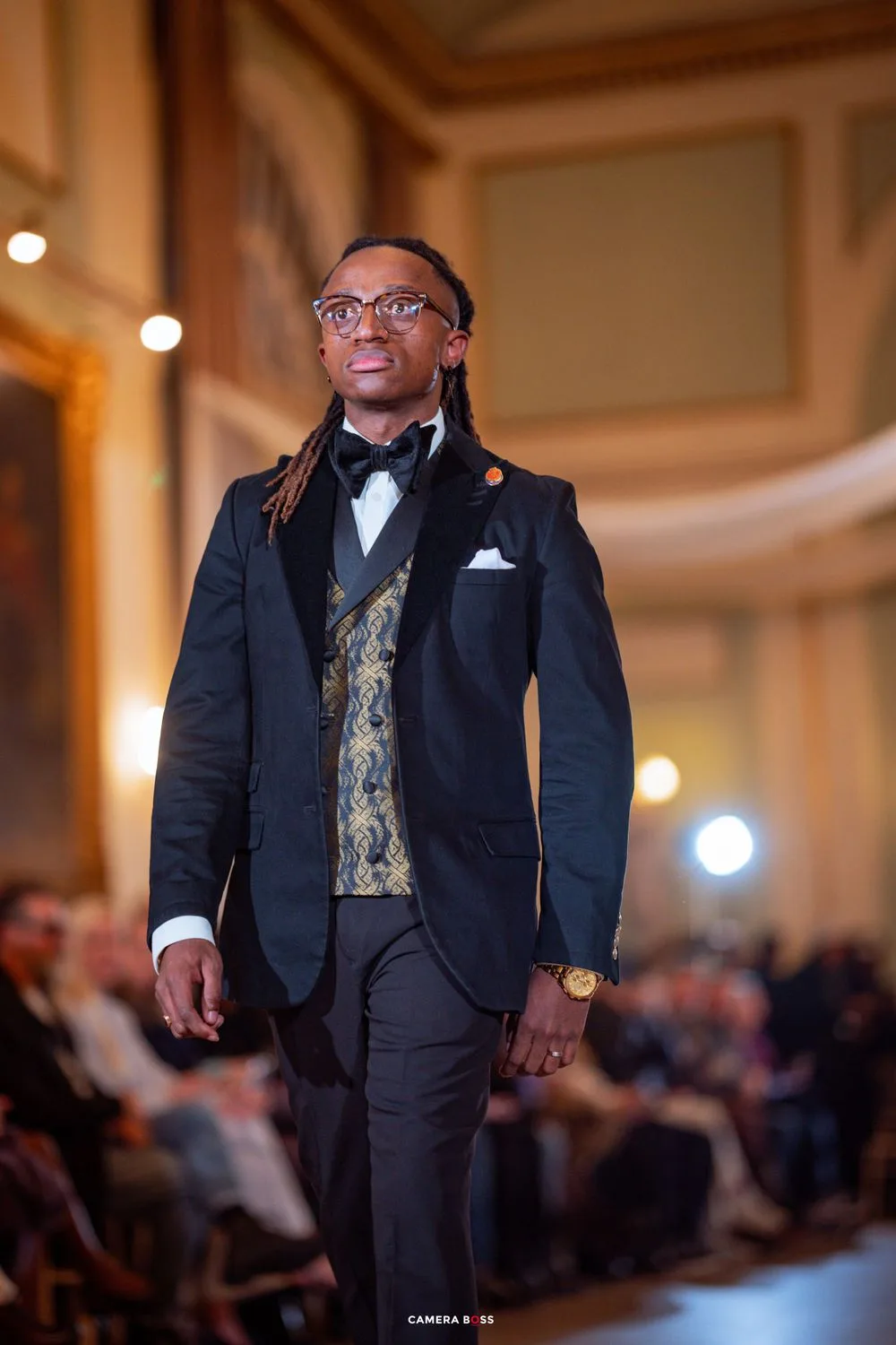 Model walks down the runway wearing black suit with ornate patterned vest and bowtie at formal fashion show.