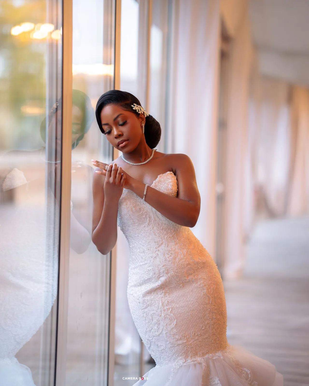 A bride in an elegant white mermaid wedding gown stands by a window in soft natural light, checking her phone.
