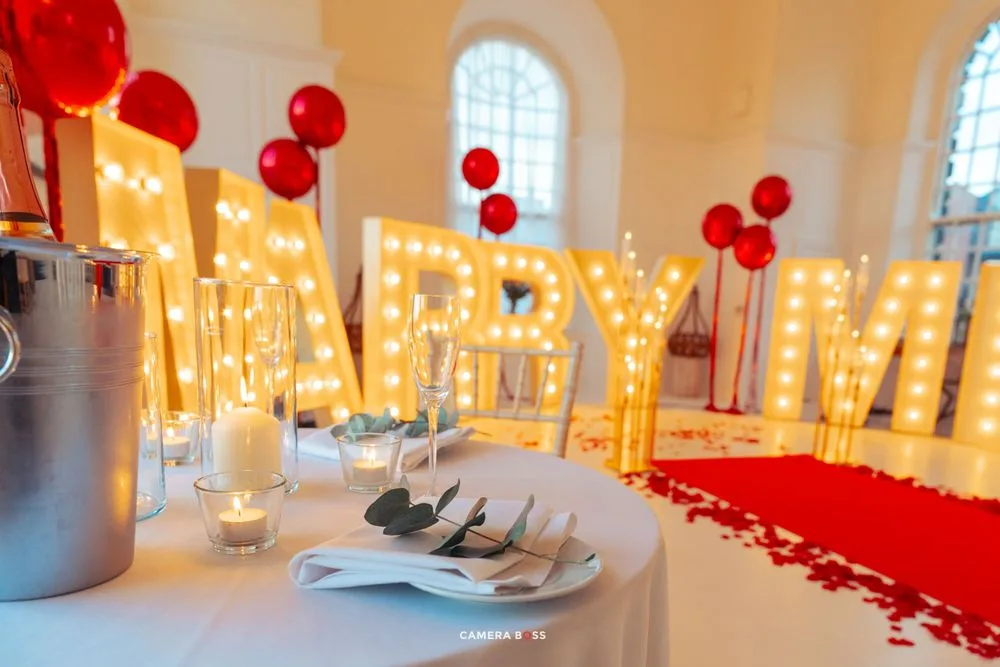 Romantic party setup with illuminated MARRY ME letters, red balloons, and rose petals along a white table.