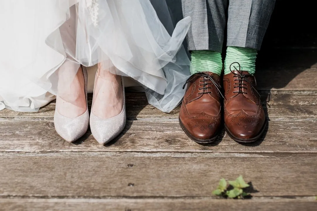 Wedding couple shows off contrasting footwear with white bridal heels and brown leather dress shoes with green socks on wooden floor.