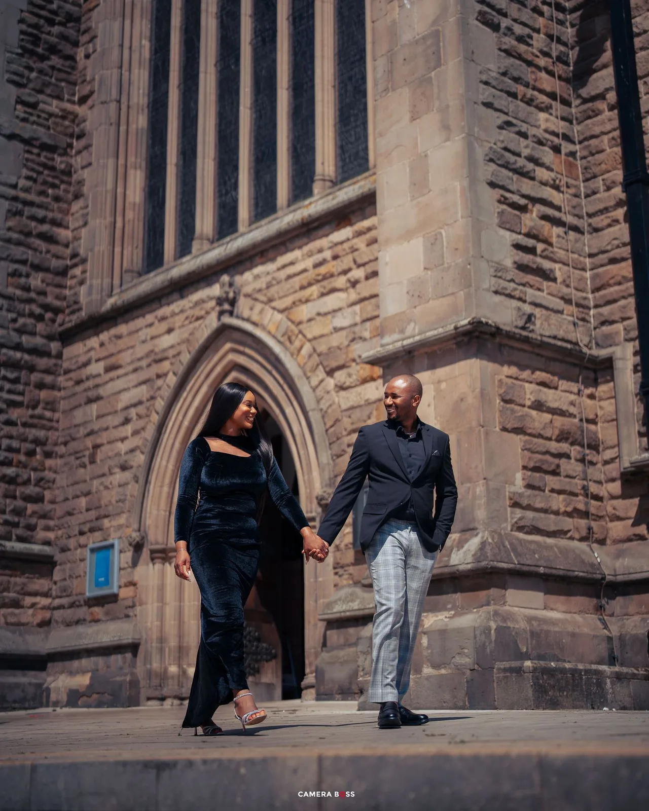Elegant couple holding hands walking in front of a historic stone church, dressed in formal black attire.
