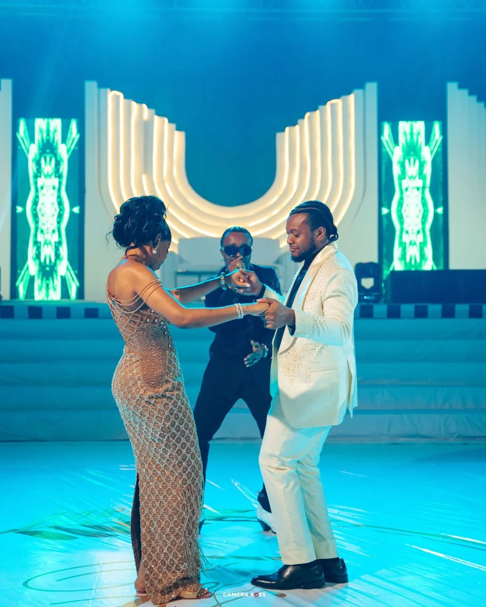 Three people dancing and performing on an illuminated blue stage with glowing white archways and neon accents in the background.
