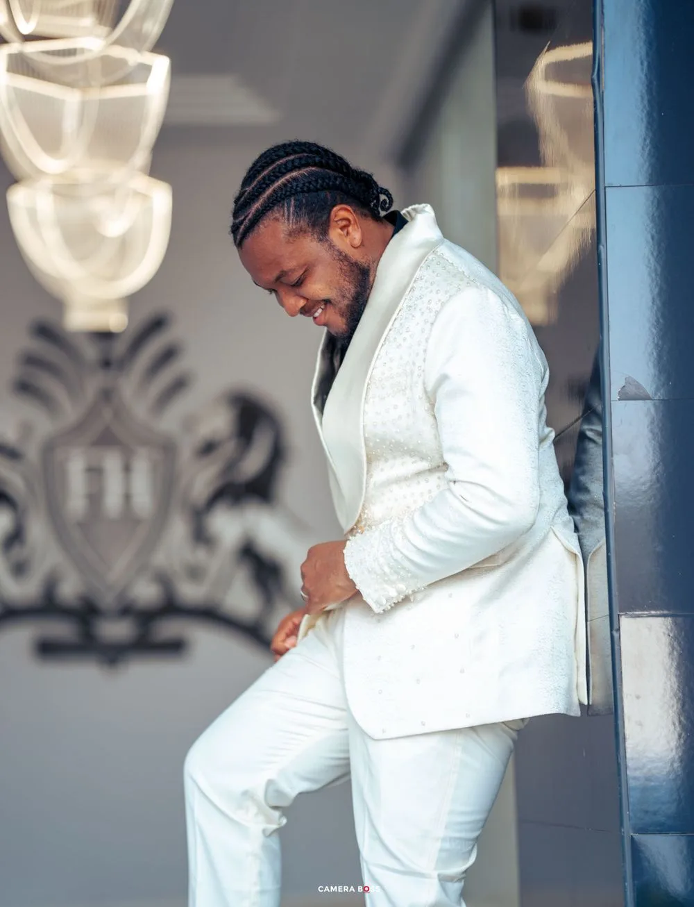 Someone in a stylish white suit smiles while posing against a decorative wall and blue column.