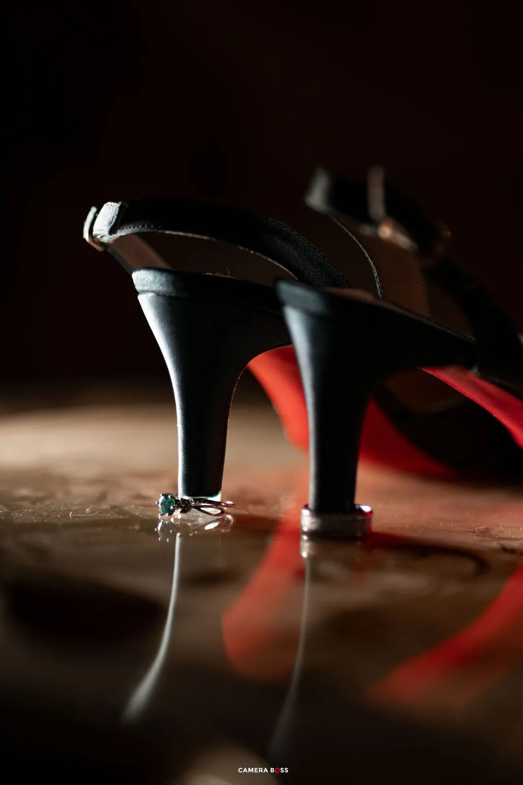 Close-up of elegant black high heel shoes with red soles and a diamond ring perched on the heel in moody lighting.
