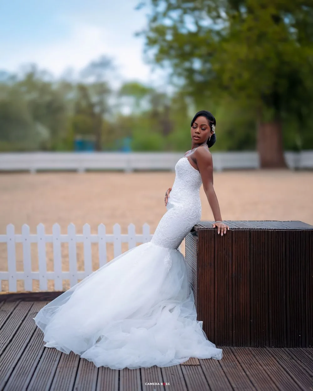 A person in an elegant white mermaid wedding dress poses gracefully on a wooden deck near a white picket fence.
