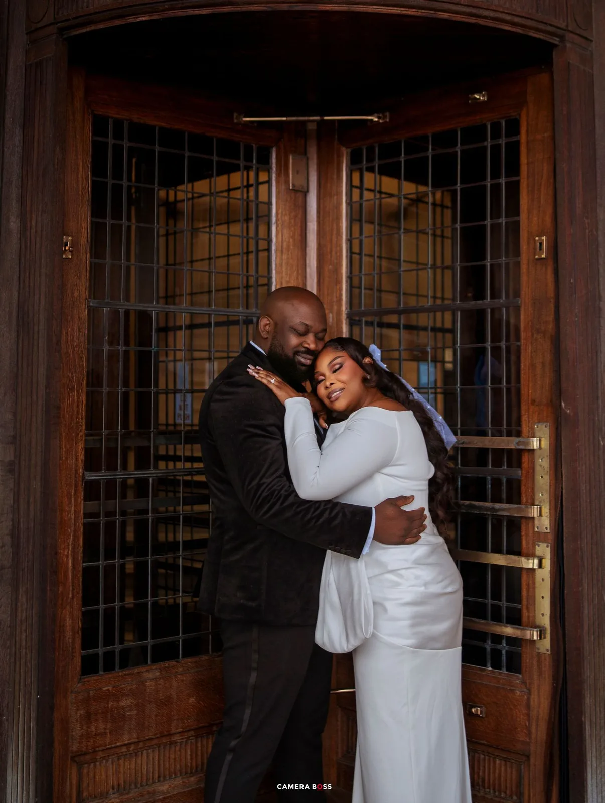 A couple in wedding attire embraces tenderly before ornate wooden doors, the bride in white and groom in a dark suit.