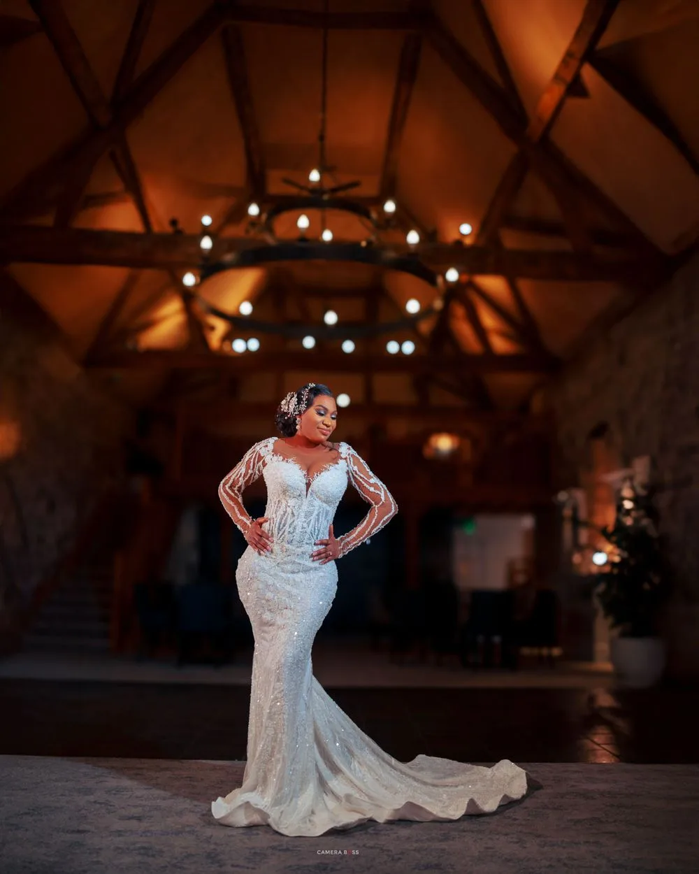 A beautiful long-sleeved lace wedding dress is showcased under dramatic wooden beams and warm lighting at a rustic venue.