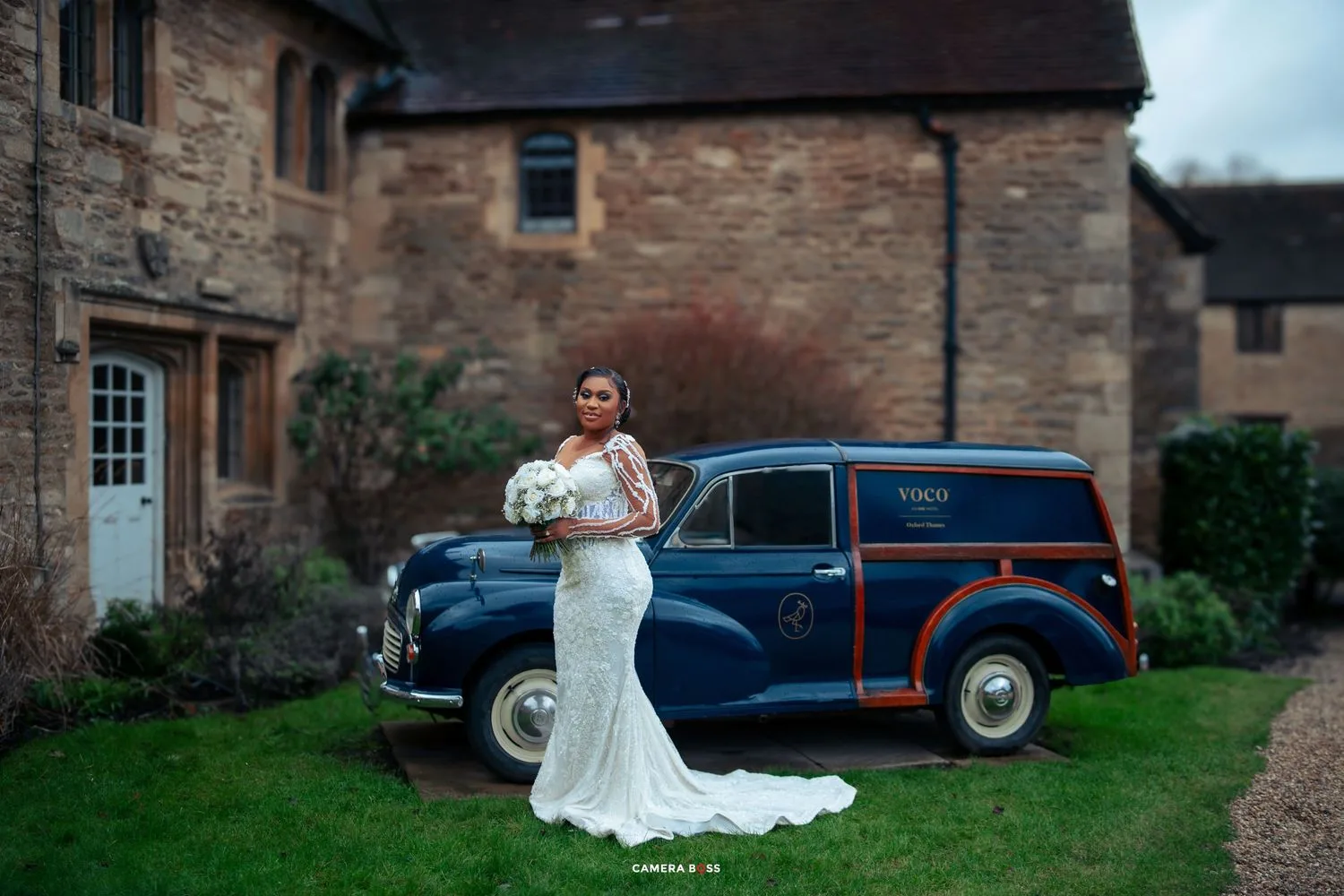 White lace wedding dress photographed next to classic blue vintage car at rustic stone cottage.