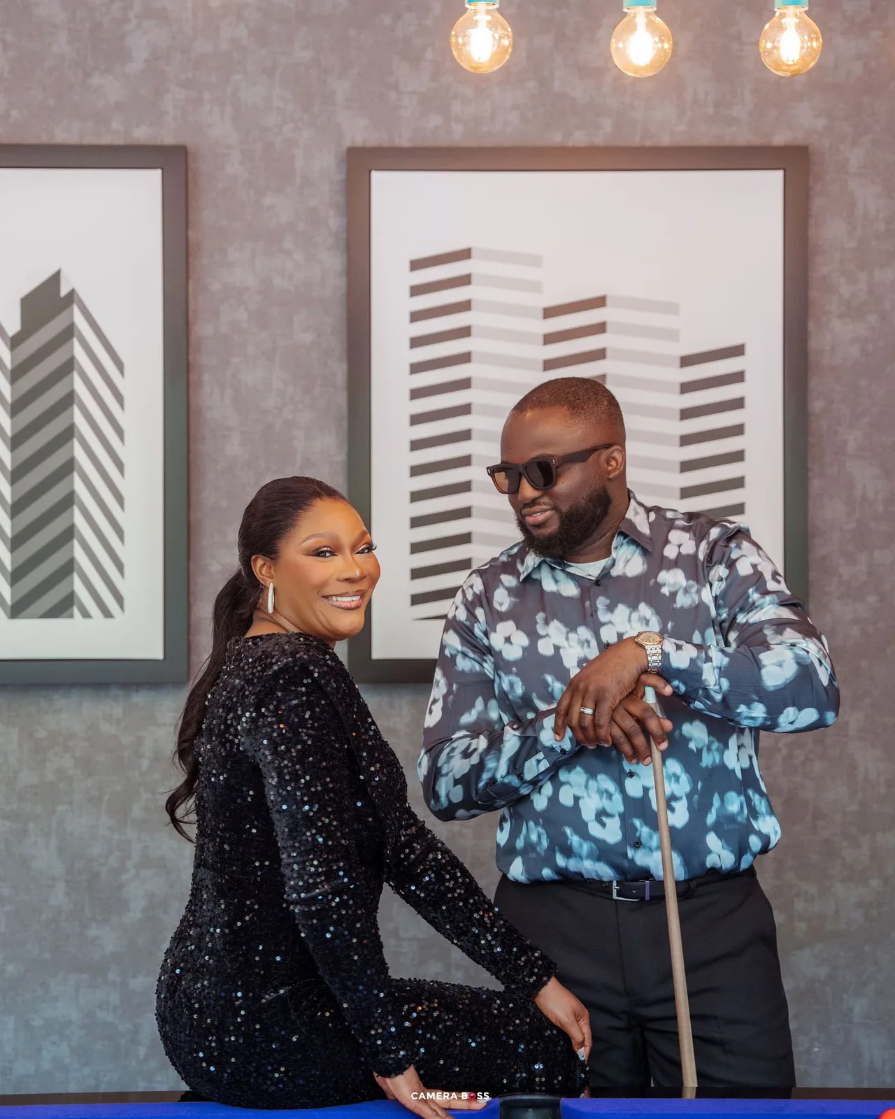 Two people in stylish attire pose and smile inside a room with modern art and pendant lights.