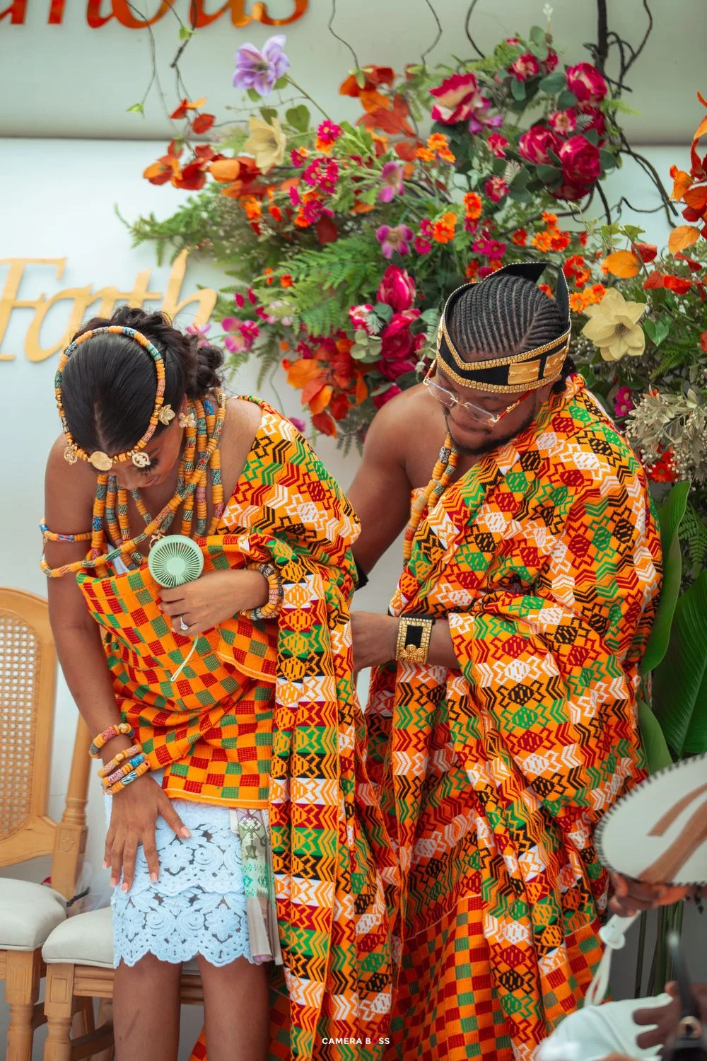 Traditional African wedding celebration with vibrant orange and red kente cloth attire and floral decorations.