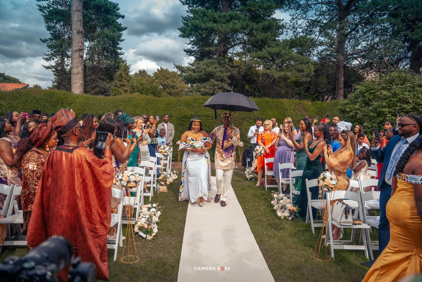 Guests gather at an outdoor wedding ceremony with white chairs on a garden path surrounded by greenery and trees.