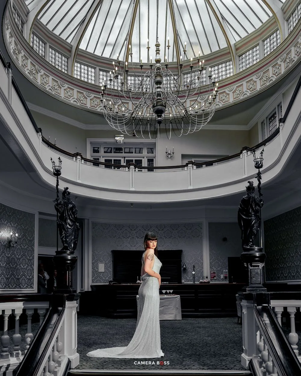 Elegant figure in white gown stands beneath ornate domed ceiling with chandelier in grand historic building interior.