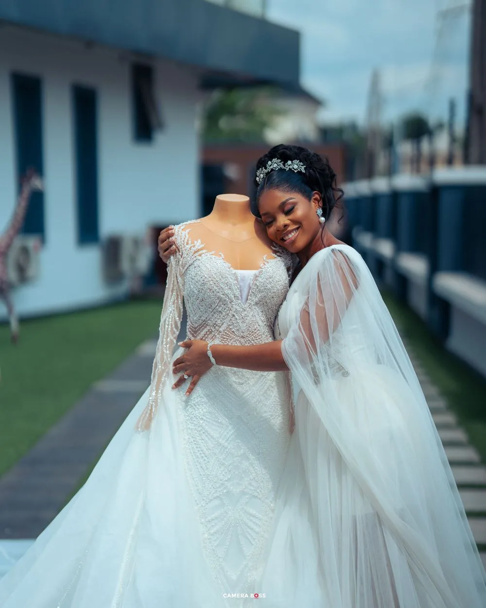 Person in a white wedding gown cape and tiara embraces a dress form mannequin displaying an elegant lace wedding dress.