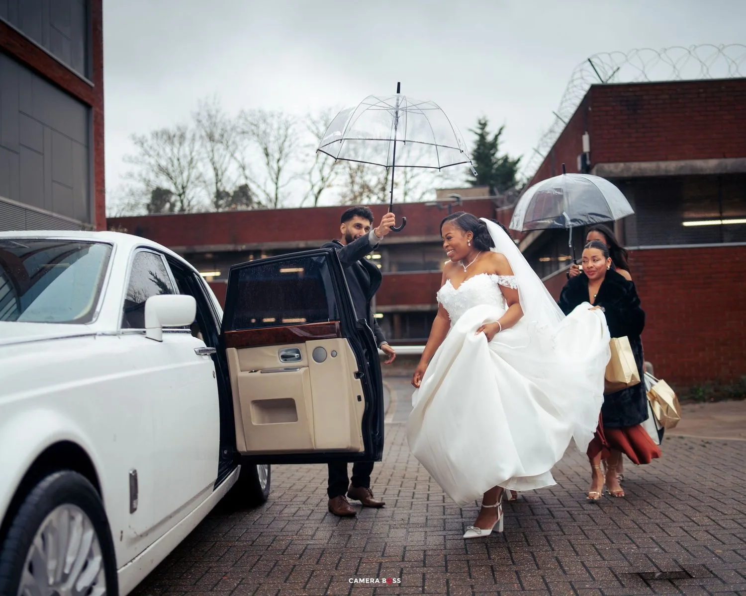 A group exits a white luxury car on a rainy day, with someone in a white dress being helped with an umbrella.