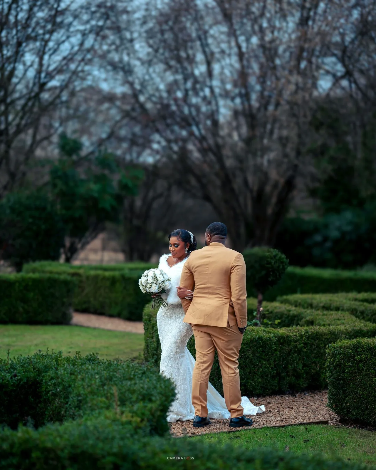 A couple in wedding attire stands on a garden path surrounded by manicured hedges and trees at dusk.