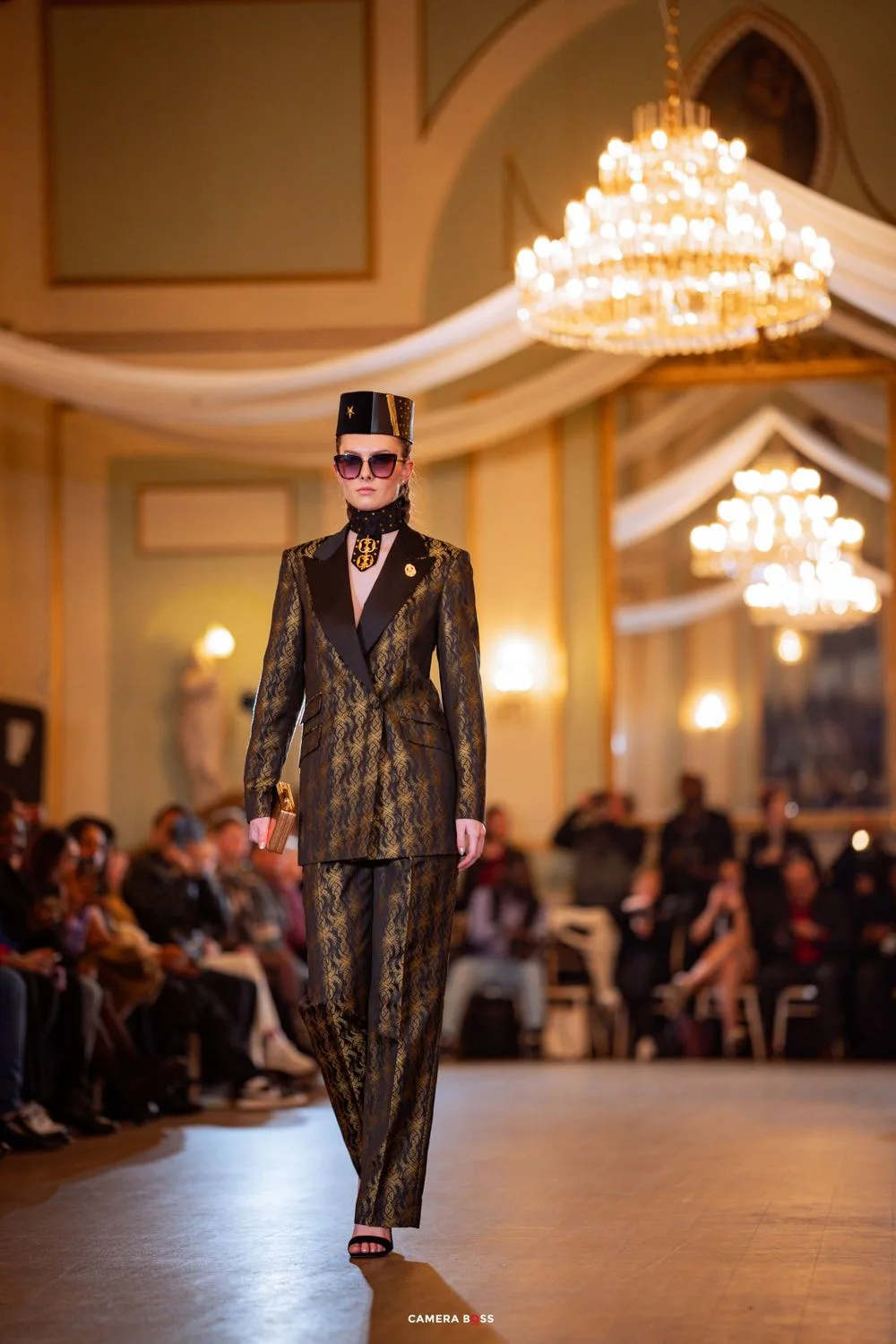 A model walks the runway in a metallic gold pantsuit and sunglasses at an elegant ballroom fashion show.