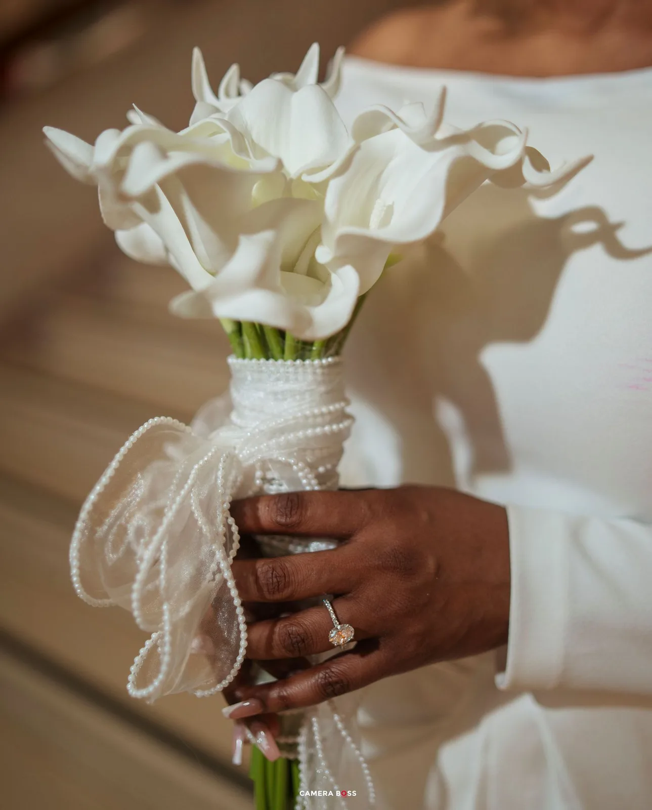 Bride holding white calla lily bouquet wrapped in pearl strands, wearing diamond ring, in white wedding dress.