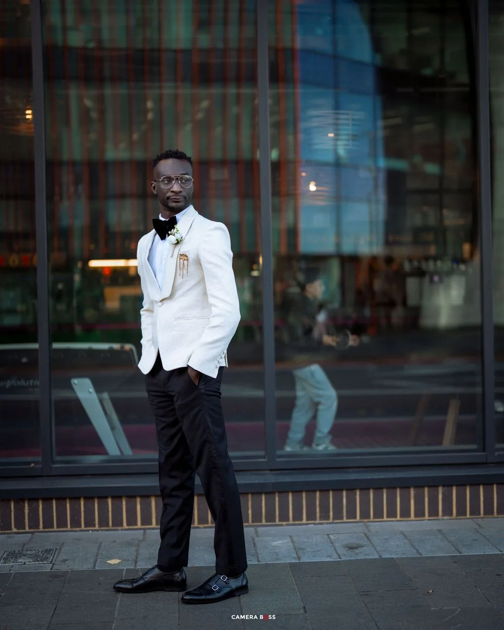 Person in crisp white tuxedo jacket and black pants poses outdoors near modern building windows.