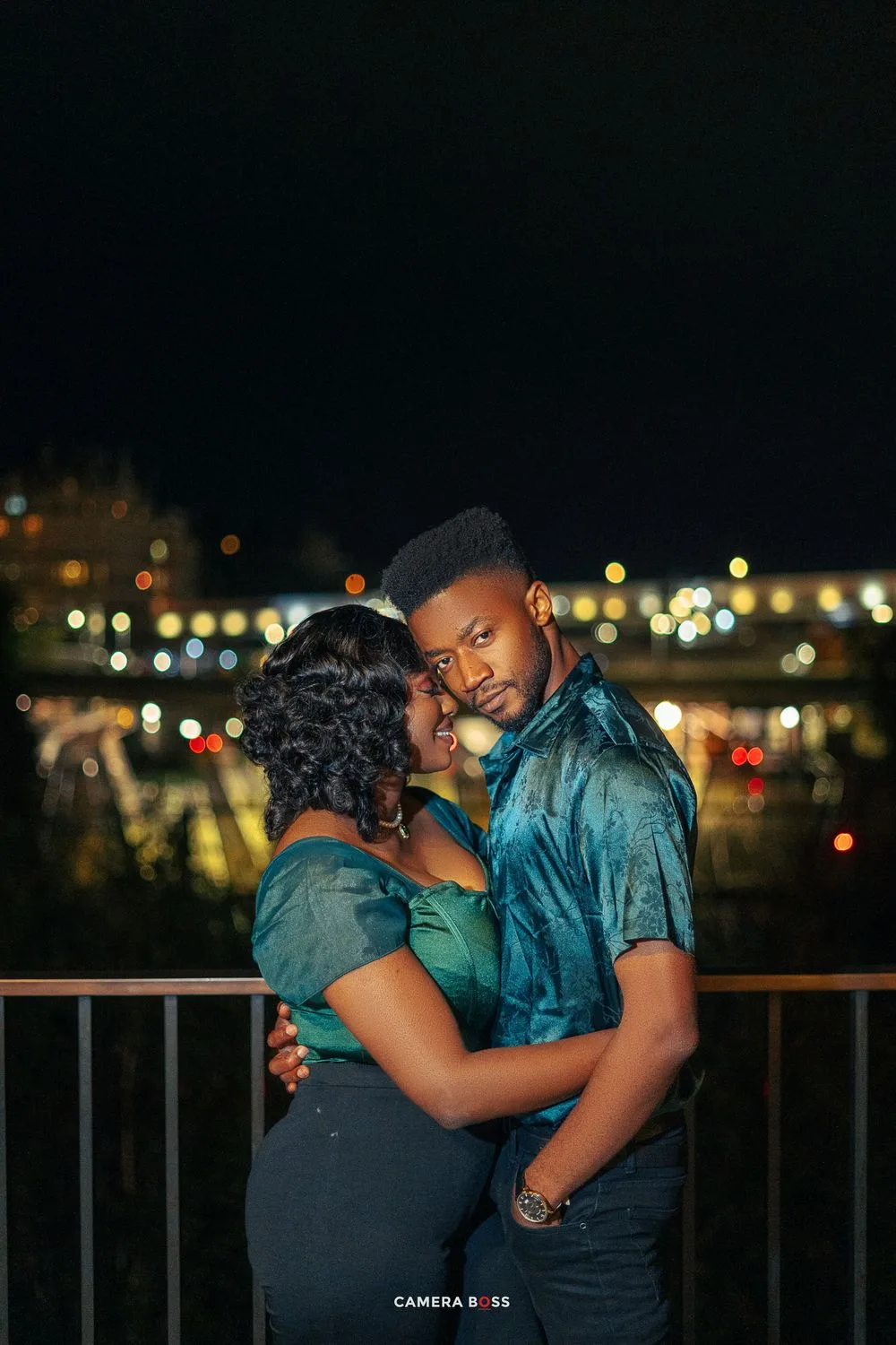 Romantic couple shares intimate moment on balcony at night with city lights twinkling in background.