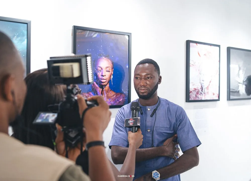 A person speaks into a microphone during a media interview as a camera operator films against a wall of framed portraits.