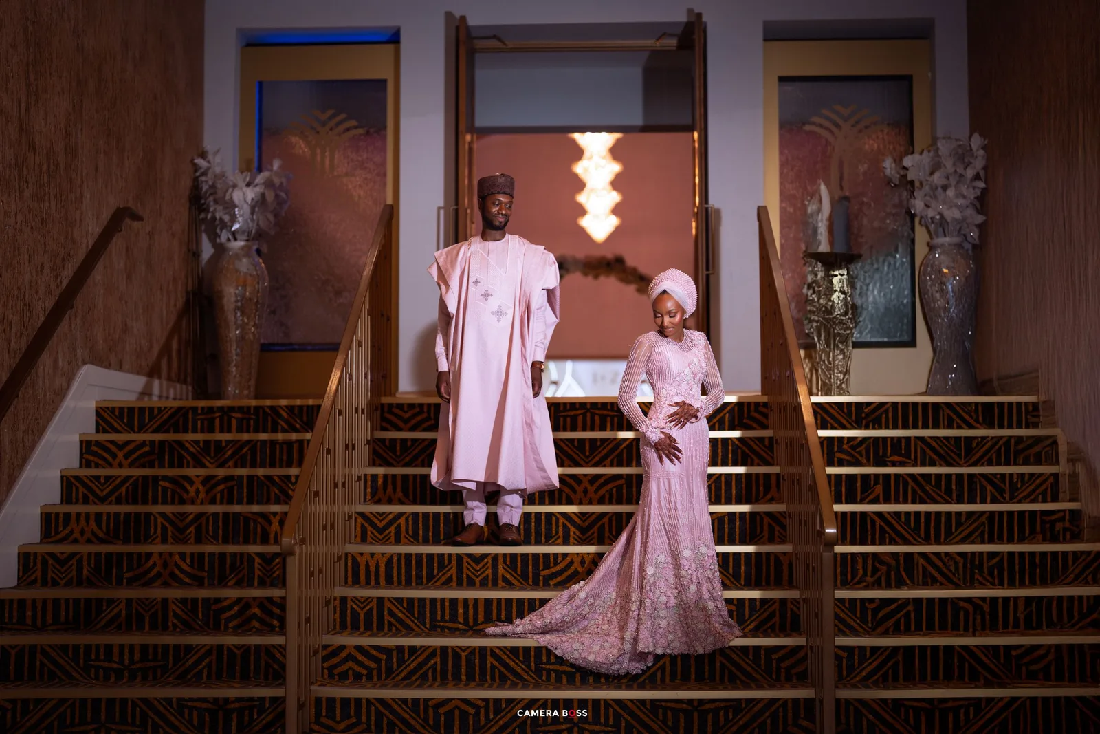 Nigerian couple in matching pink traditional attire posing elegantly on grand staircase during wedding ceremony.