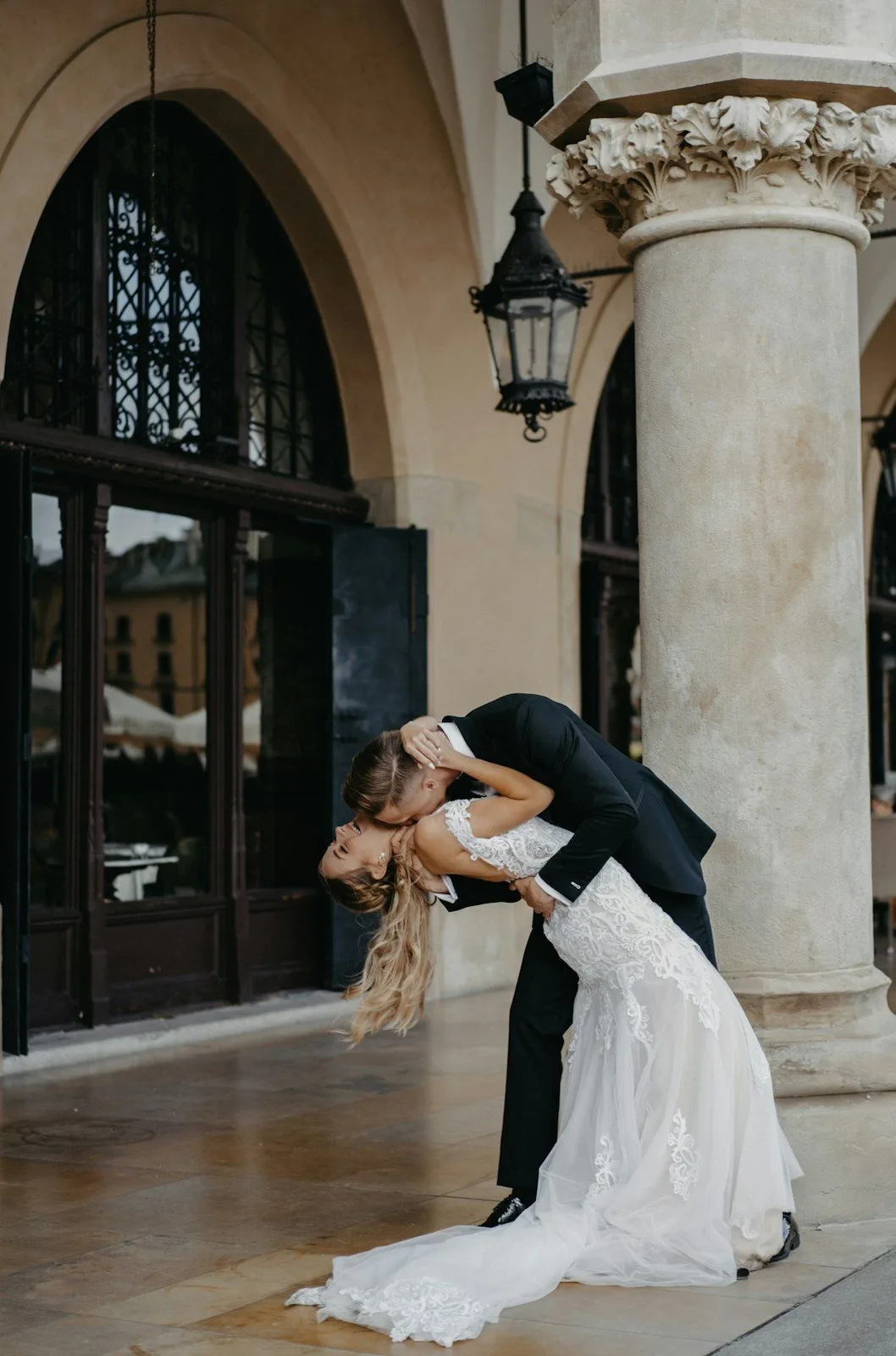 Romantic wedding couple shares a dramatic dip kiss under an ornate archway with vintage lantern and columns.