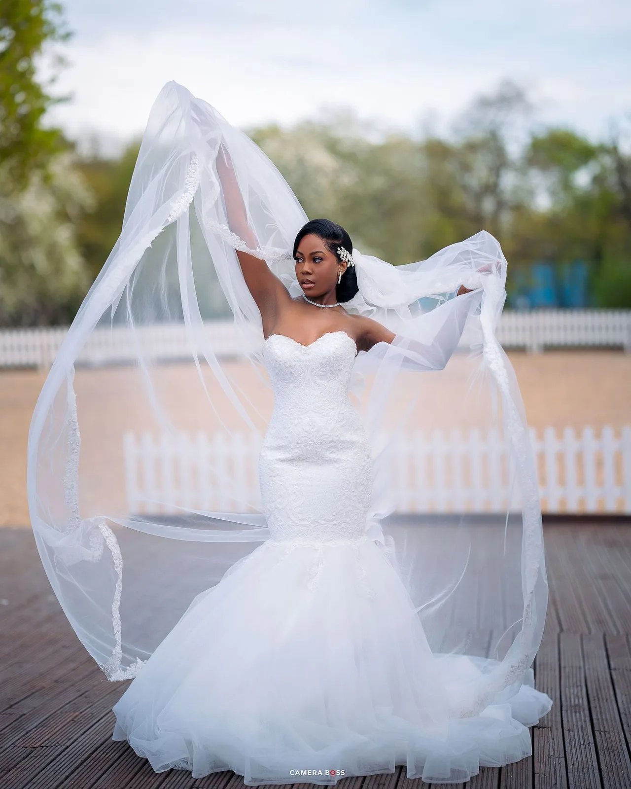 A bride in a fitted mermaid wedding gown with flowing veil stands elegantly on a wooden deck with white fence background.