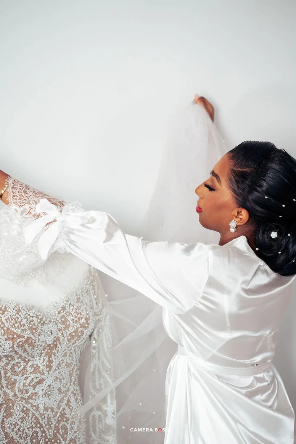 Wedding preparation scene with elegant white satin robe and delicate lace dress hanging against light wall.