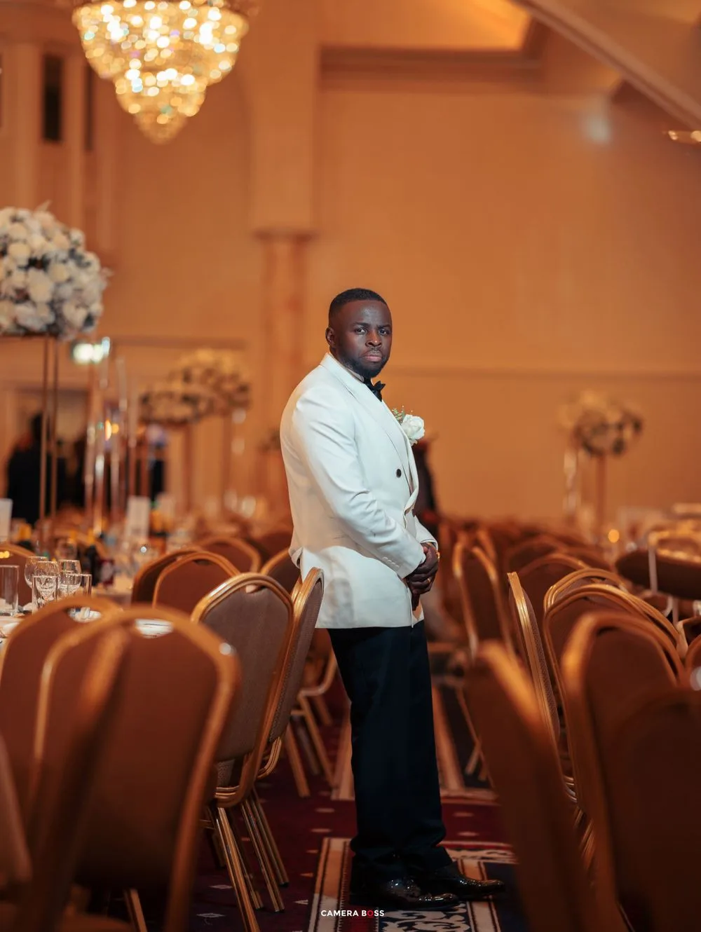 Someone in a white tuxedo jacket stands elegantly in an elegant ballroom with chandelier and banquet chairs.