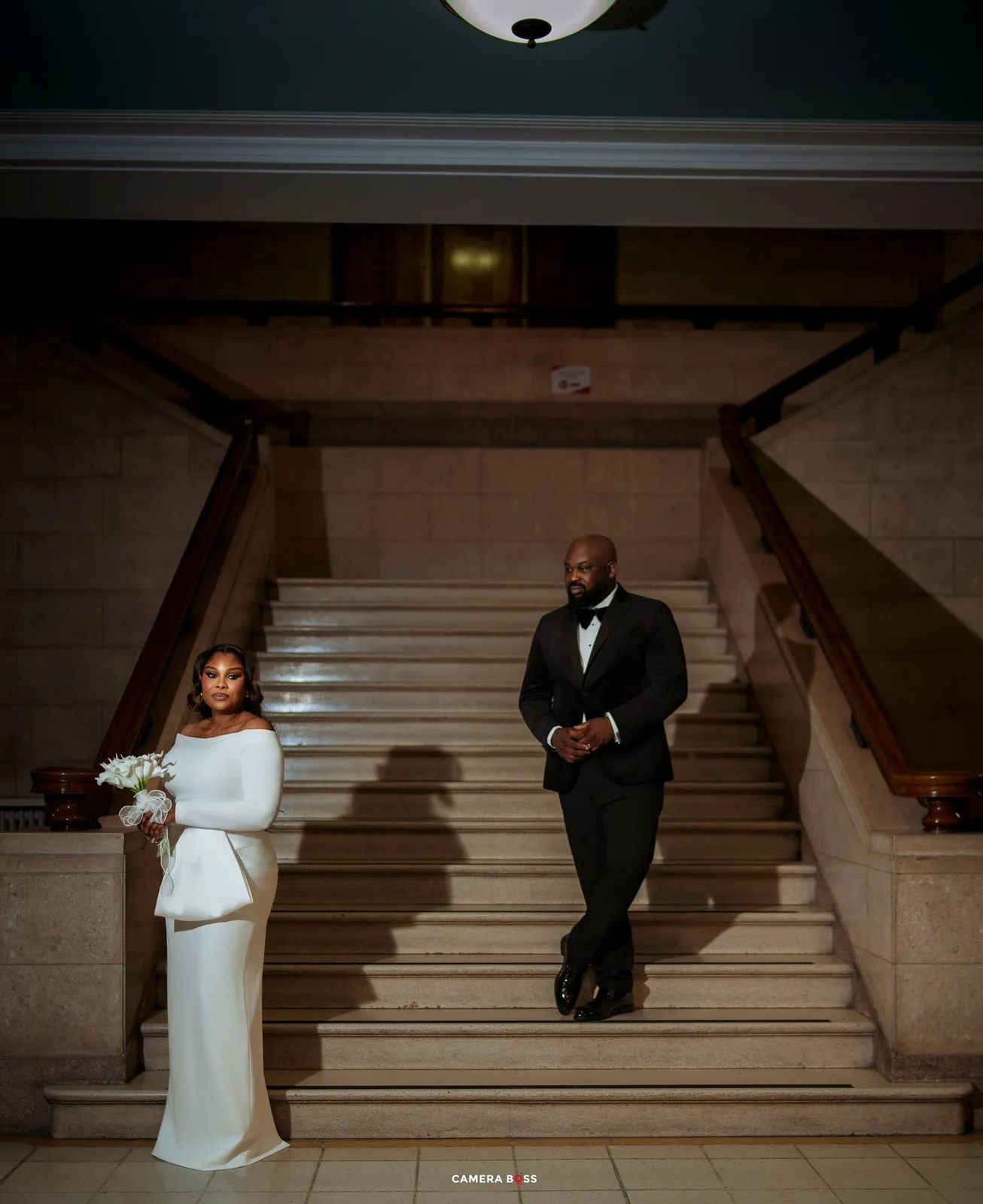 Elegant couple in wedding attire descending grand marble staircase, bride in white gown holding bouquet, groom in black tuxedo.