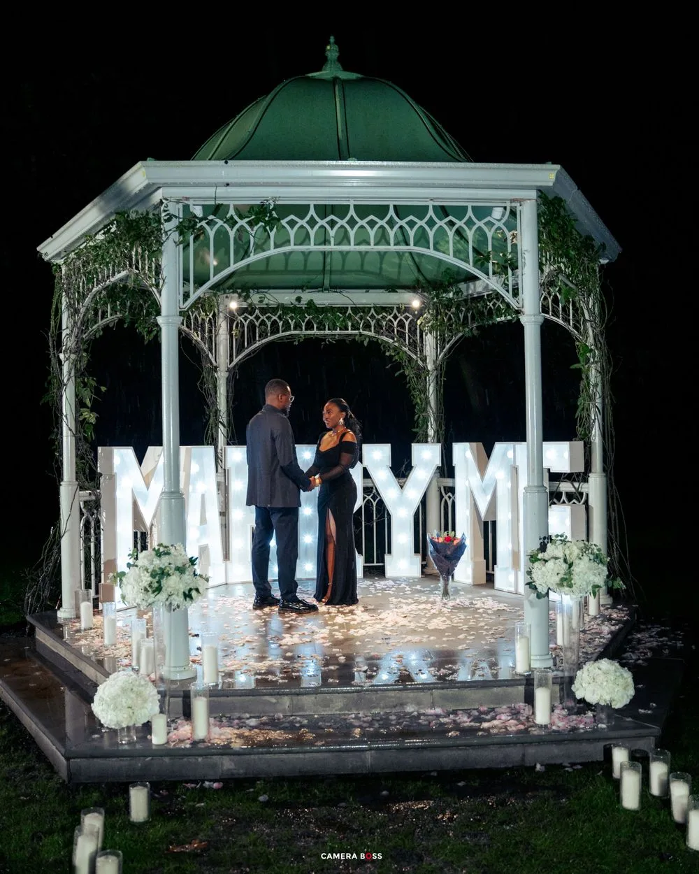 A illuminated white gazebo decorated with flowers and 'MARRY ME' letters for a romantic nighttime proposal.