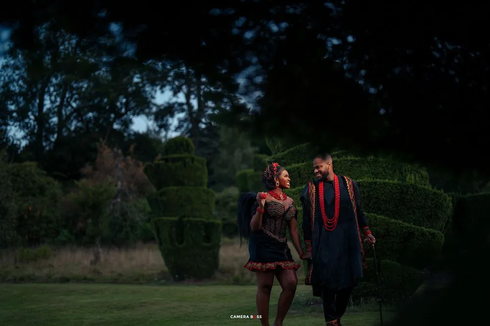 Two graduates in red and blue graduation gowns walk together in a garden with ornamental hedges at dusk.