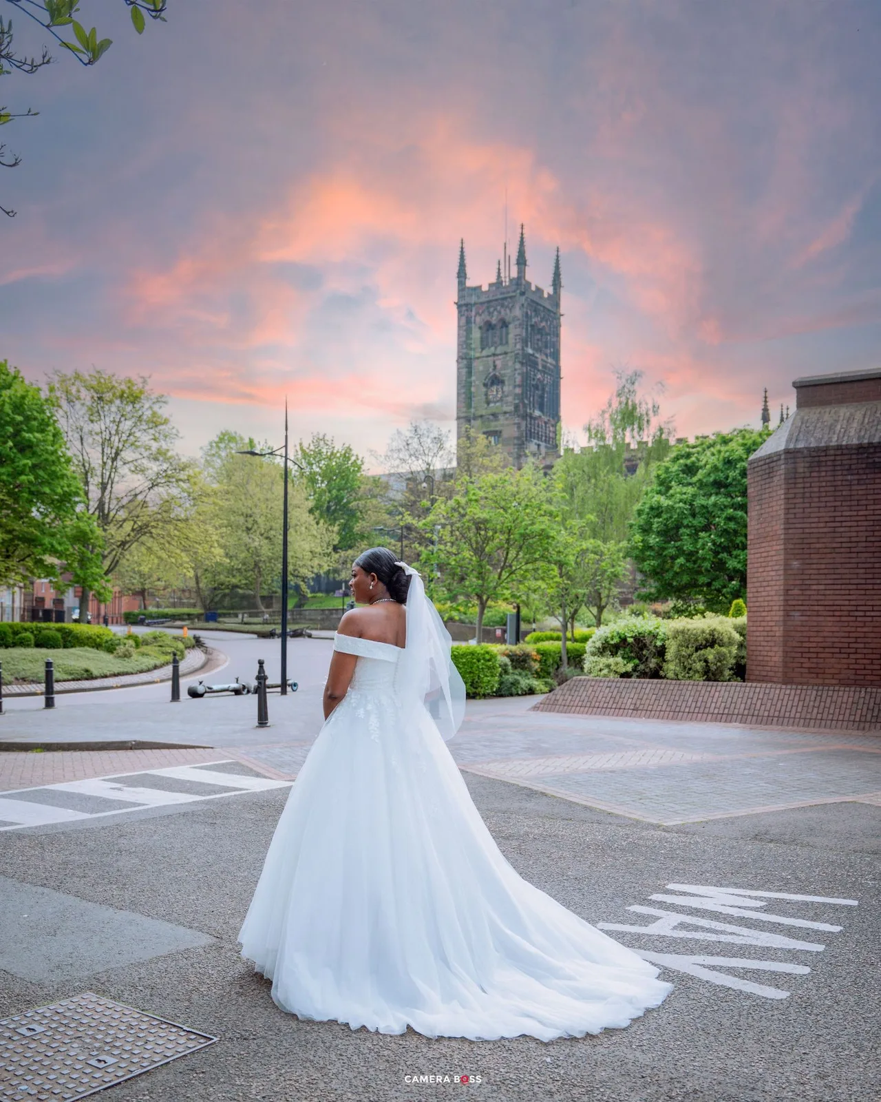 A bride in a flowing white gown and veil poses against a sunset sky with a gothic church spire in the background.