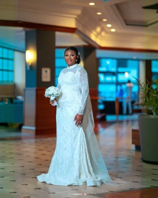 Beautiful bride in a white long-sleeve lace wedding gown and veil poses with bouquet in an elegant hotel lobby setting.