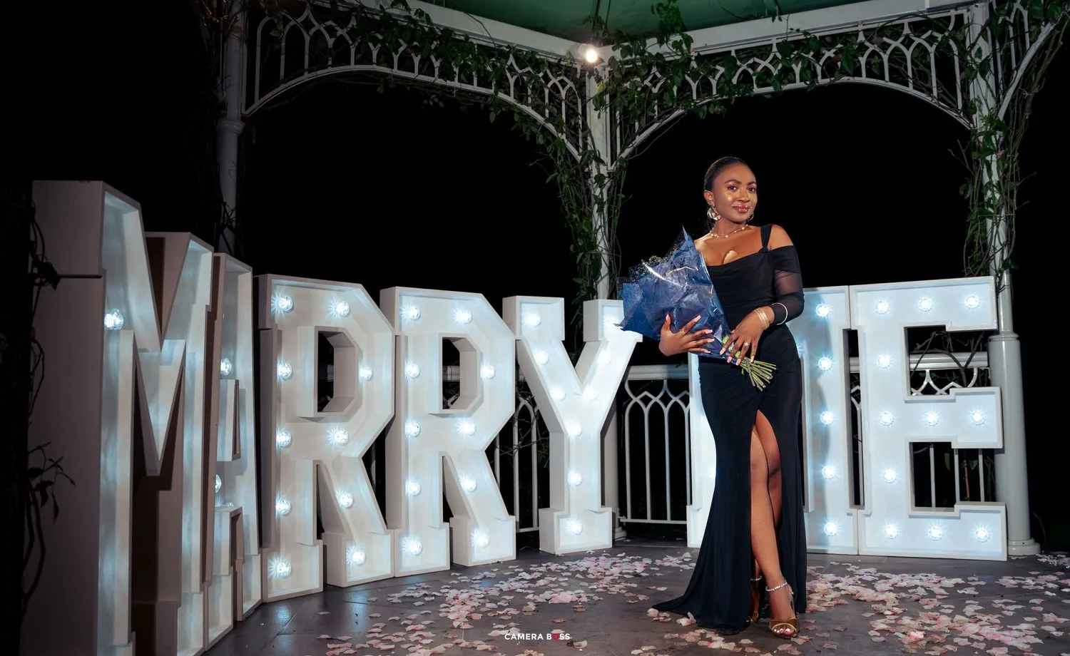 A person stands elegantly in front of illuminated MARRY ME letters at night under an ornate gazebo.